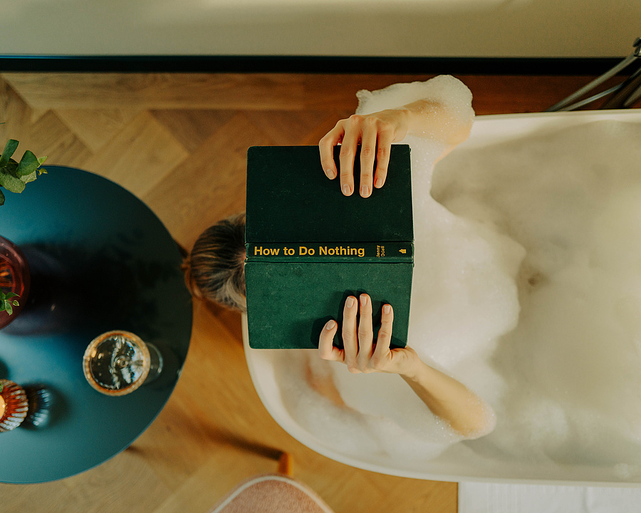Frau in einer schaumigen Badewanne liest ein Buch mit dem Titel "How to Do Nothing", mit einem blauen Beistelltisch, auf dem ein Glas Wein und Blumen stehen