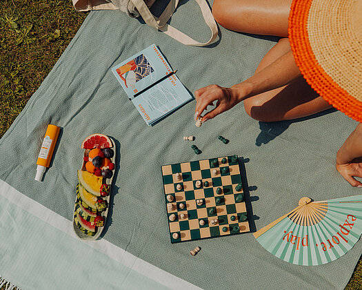 Frau mit großem Strohhut sitzt auf grauer Decke auf der Wiese und spielt Schach. Um sie herum liegen ein Fächer, eine Obstplatte, Sonnencreme und ein Buch.