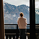 Frau in grauer Hose und beigem Pullover steht auf dem Balkon und blickt in die winterliche Berglandschaft mit See. Aufnahme aus dem Zimmer.
