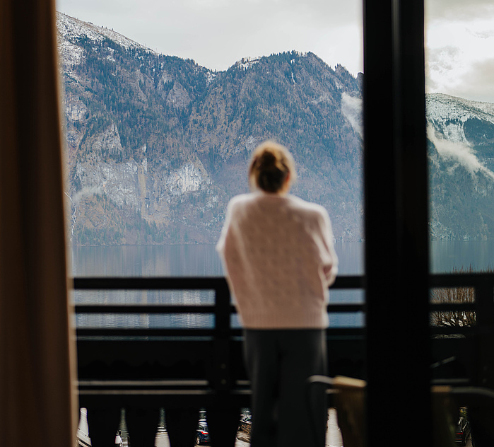Frau in grauer Hose und beigem Pullover steht auf dem Balkon und blickt in die winterliche Berglandschaft mit See. Aufnahme aus dem Zimmer.