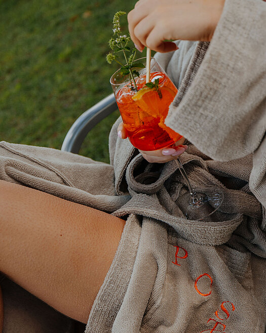 Frau im Bademantel sitzt draußen auf einem Stuhl in der Wiese und hält einen orangen Cocktail in der Hand.