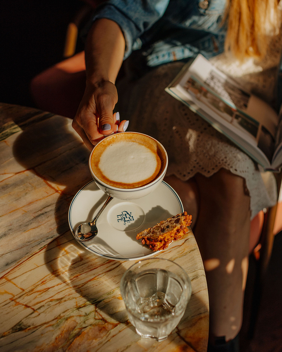 Frau im weißen Kleid und Jeansjacke sitzt entspannt im Restaurant bei einer Tasse Kaffee