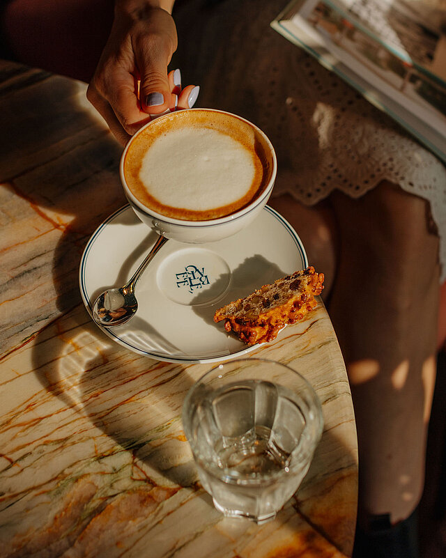 Frau im weißen Kleid und Jeansjacke sitzt entspannt im Restaurant bei einer Tasse Kaffee