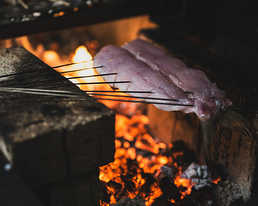 Frisch gegrillter Fisch aufgespießt über offenem Feuer, knisternde Flammen im Hintergrund