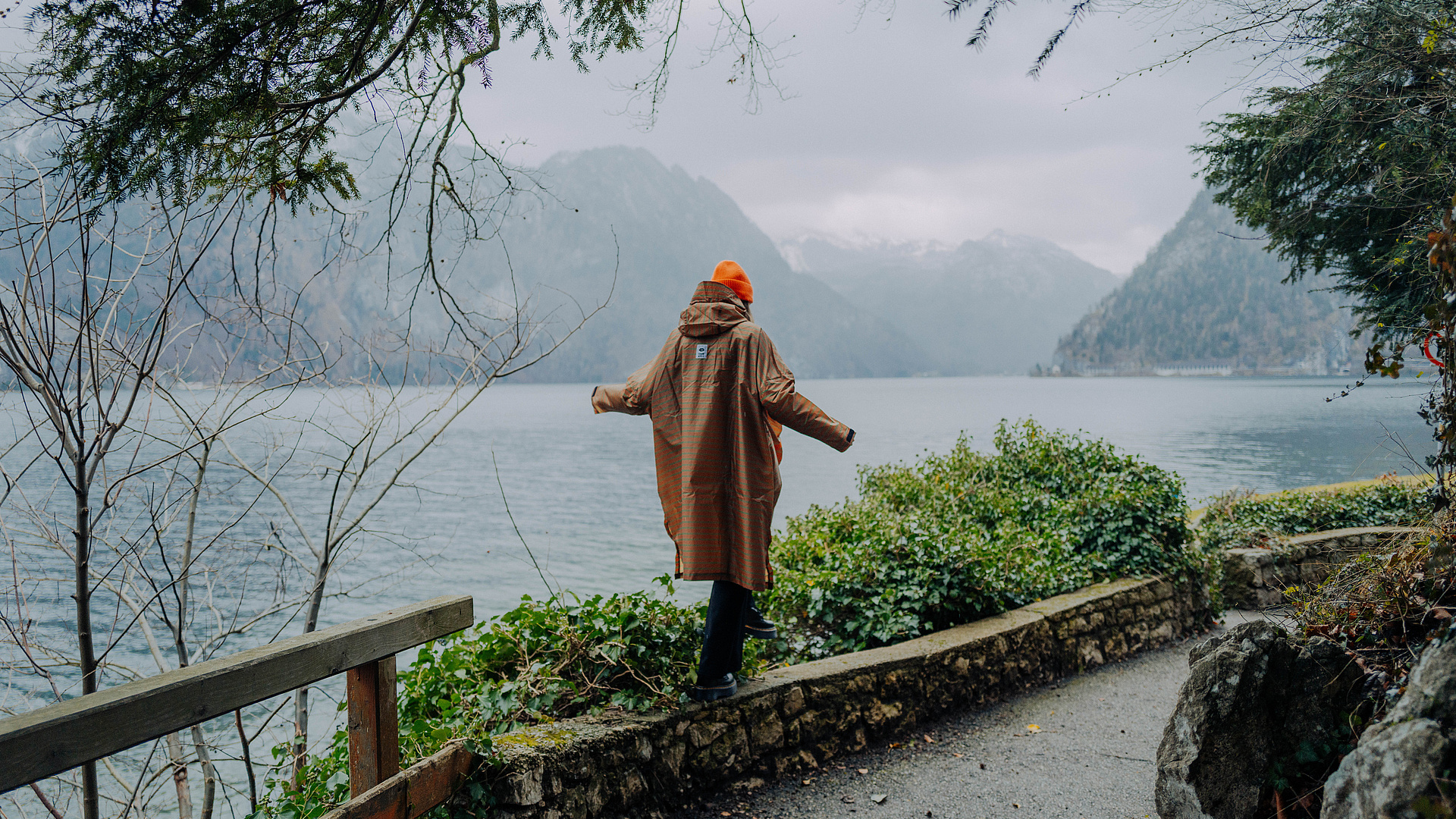 Person im braunen Regencape balanciert auf einer Steinmauer im Regen, im Hintergrund die Berge und der Traunsee