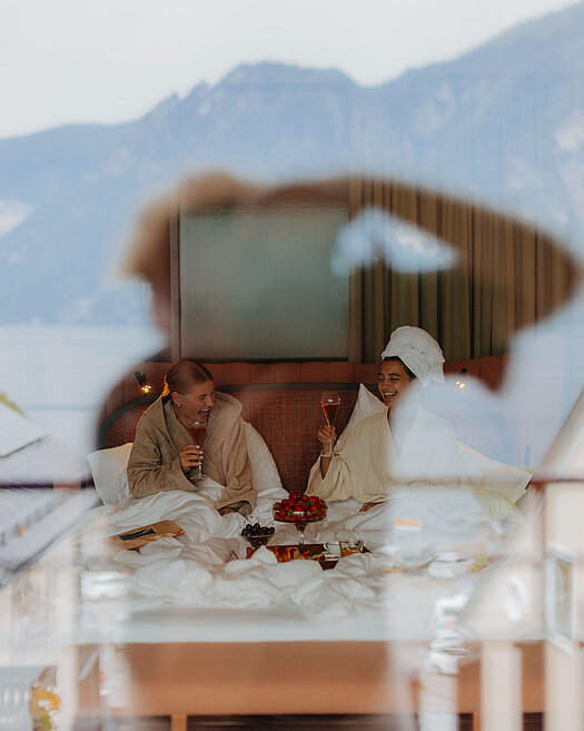 Zwei Frauen im Bademantel sitzen lachend im Bett mit einem Glas Sekt in der Hand. Vor ihnen steht ein Tablett mit süßen Köstlichkeiten. In der Glasreflexion sind der Fotograf, der See und die Berge zu sehen.