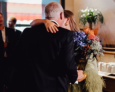 Mann im schwarzen Anzug mit einem Blumenstrauß umarmt eine Frau in einem ausgefranzten hellgrünen Kleid im Restaurant Beletage.