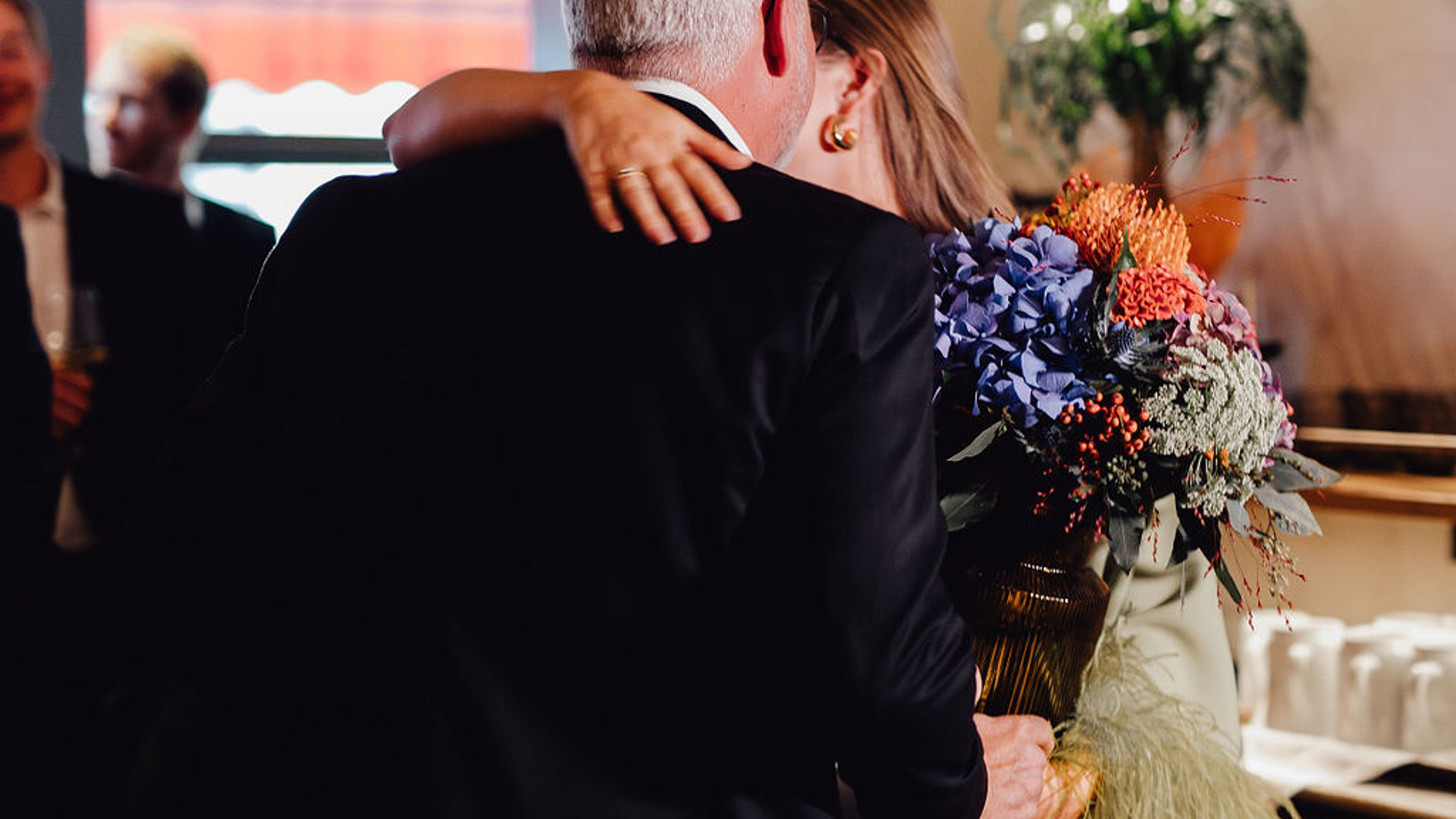 Mann im schwarzen Anzug mit einem Blumenstrauß umarmt eine Frau in einem ausgefranzten hellgrünen Kleid im Restaurant Beletage.