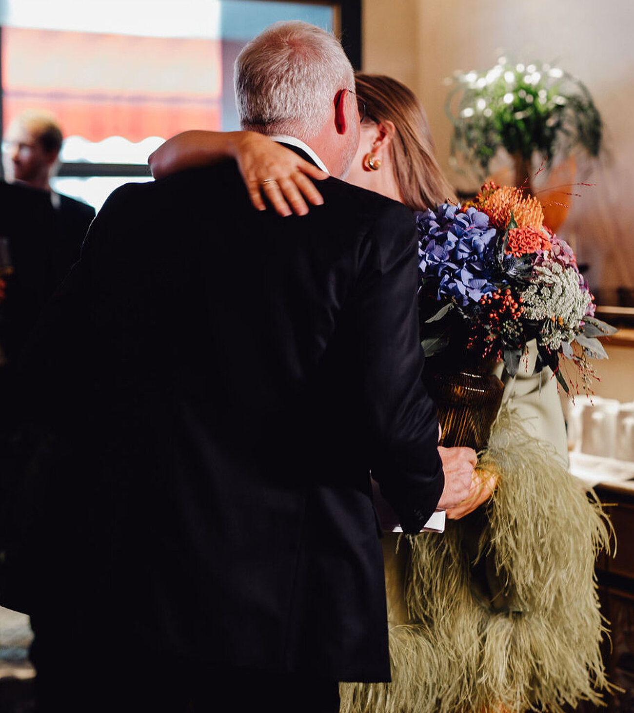 Mann im schwarzen Anzug mit einem Blumenstrauß umarmt eine Frau in einem ausgefranzten hellgrünen Kleid im Restaurant Beletage.