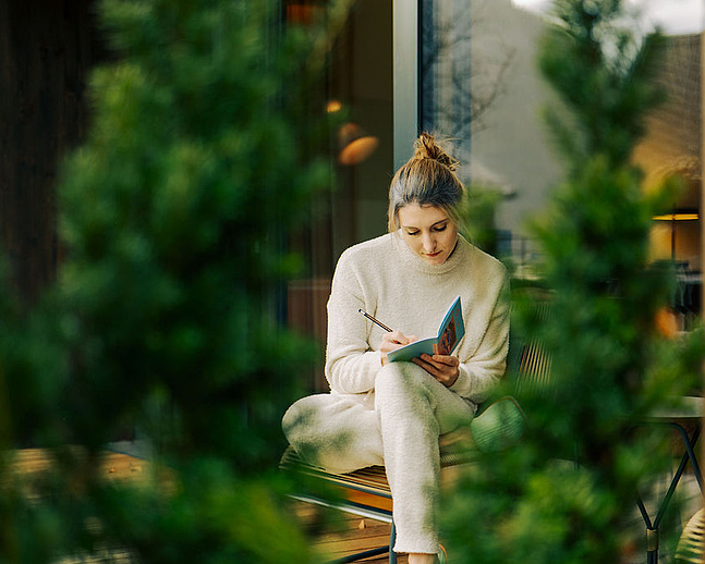 Frau in beiger Kleidung schreibt in ein Notizbuch auf der Terrasse, umgeben von Natur. Im Hintergrund eine moderne Fensterfront, im Vordergrund grüne Äste.
