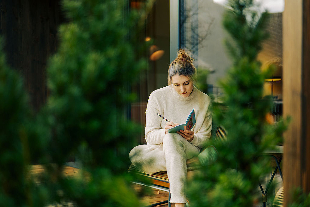Frau in beiger Kleidung schreibt in ein Notizbuch auf der Terrasse, umgeben von Natur. Im Hintergrund eine moderne Fensterfront, im Vordergrund grüne Äste.