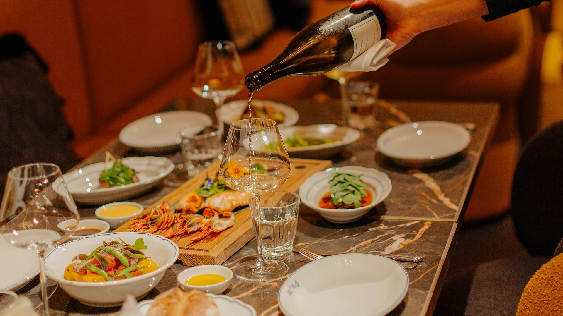 Einladender Restaurantbereich mit Gästen, während Wein in Gläser eingeschenkt wird. Verschiedene Speisen sind auf dem Tisch angerichtet.