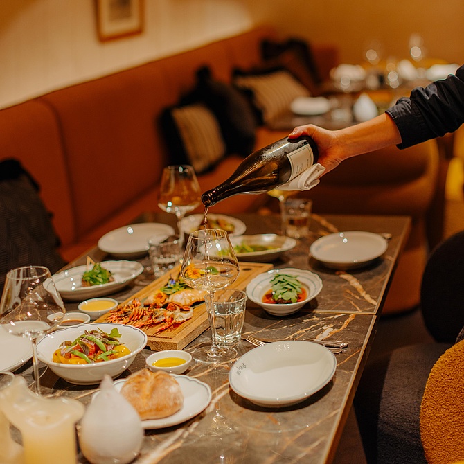 Einladender Restaurantbereich mit Gästen, während Wein in Gläser eingeschenkt wird. Verschiedene Speisen sind auf dem Tisch angerichtet.