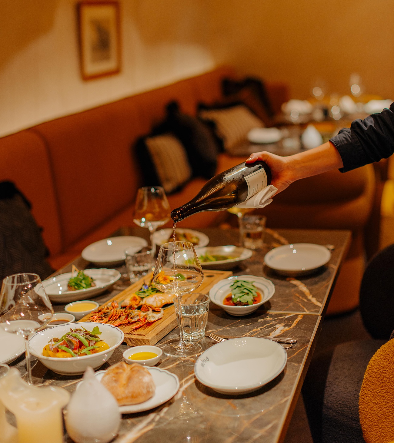Einladender Restaurantbereich mit Gästen, während Wein in Gläser eingeschenkt wird. Verschiedene Speisen sind auf dem Tisch angerichtet.