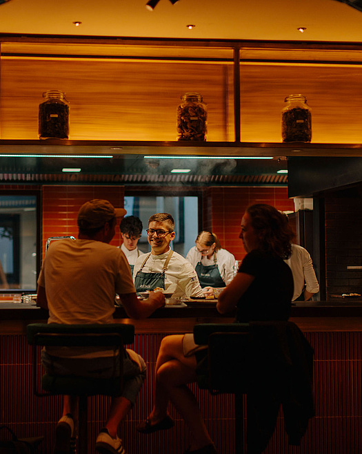 Offene Küche im Restaurant Beletage mit Köchen bei der Zubereitung. Gäste sitzen am Counter und beobachten das Geschehen