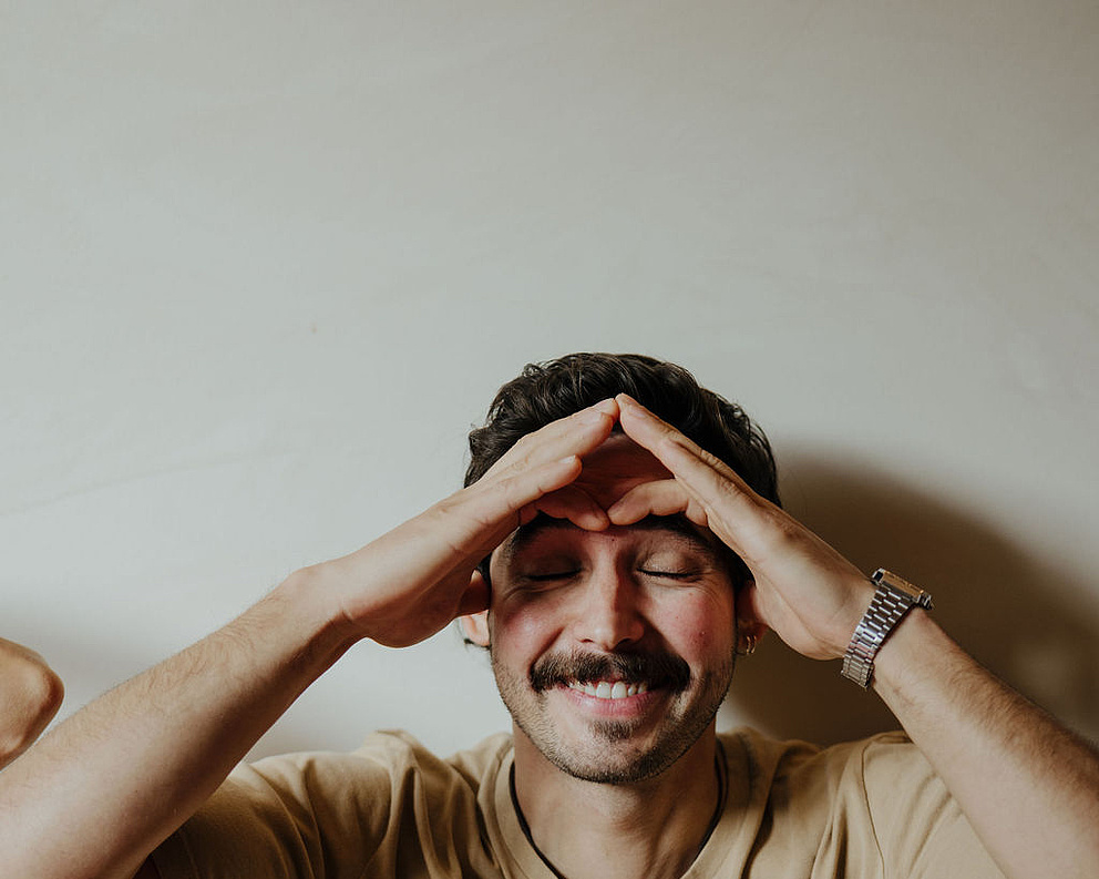 Mann in beigem T-Shirt und mit Uhr vor einer weißen Wand, der an einer Face Yoga-Session teilnimmt