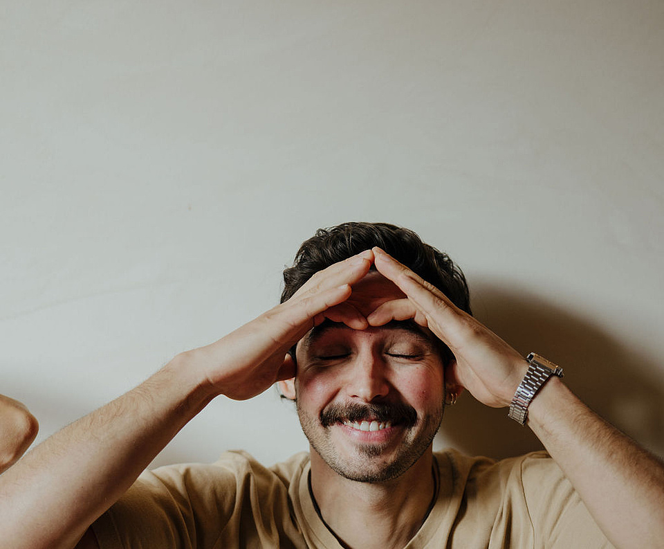 Mann in beigem T-Shirt und mit Uhr vor einer weißen Wand, der an einer Face Yoga-Session teilnimmt