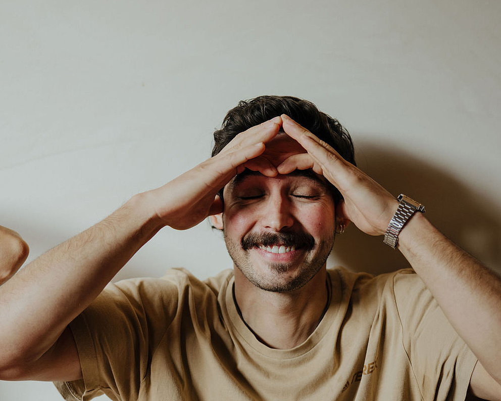 Mann in beigem T-Shirt und mit Uhr vor einer weißen Wand, der an einer Face Yoga-Session teilnimmt