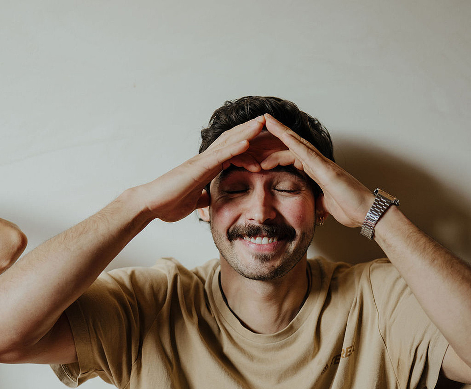 Mann in beigem T-Shirt und mit Uhr vor einer weißen Wand, der an einer Face Yoga-Session teilnimmt