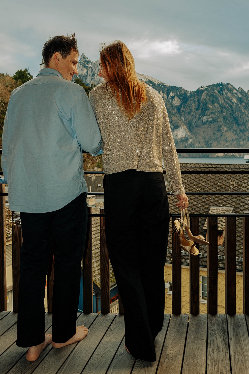 Paar auf Balkon, Mann in blauem Hemd und schwarzer Hose, Frau in goldenem glitzernden Oberteil und schwarzer Hose, beide barfuß, Frau hält High Heels in der Hand, im Hintergrund See und Berge.