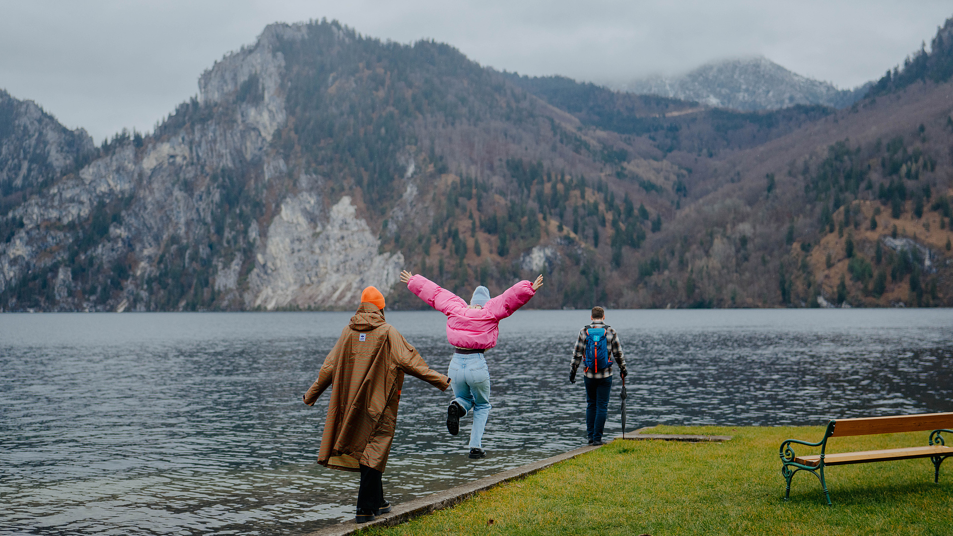 Drei Personen spazieren am Traunsee entlang, umgeben von einer Wiese mit einer Bank. Die herbstlichen Farben der Berge spiegeln sich im Wasser, während es leicht regnet.