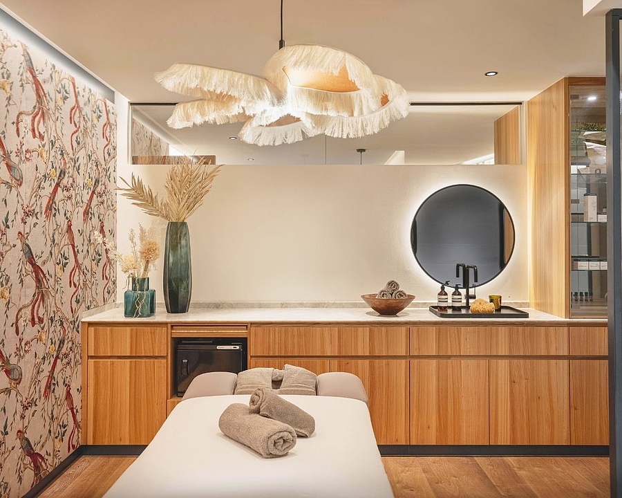 Innenansicht des Beauty- und Massagebereichs mit einer ausgefallenen Deckenlampe mit Fransen, bunten Tapeten, hellbraunen Holzmöbeln und einer Massageliege.