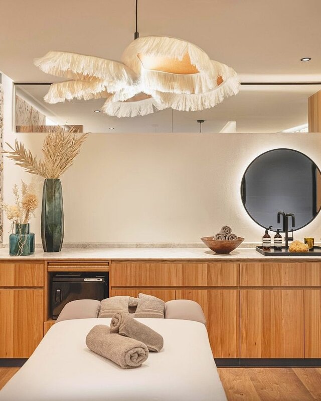 Innenansicht des Beauty- und Massagebereichs mit einer ausgefallenen Deckenlampe mit Fransen, bunten Tapeten, hellbraunen Holzmöbeln und einer Massageliege.