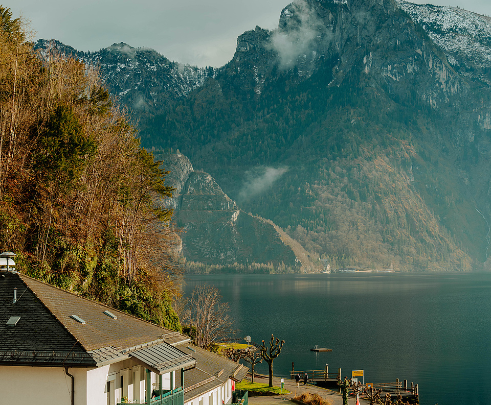 Traunsee mit der „Schlafenden Griechin“ im Hintergrund, weißes Haus mit Flaggen, Holzstege am Ufer.