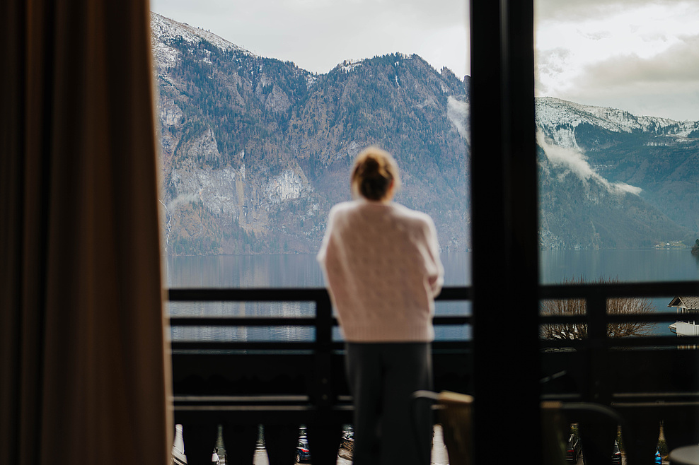 Frau in grauer Hose und beigem Pullover steht auf dem Balkon und blickt in die winterliche Berglandschaft mit See. Aufnahme aus dem Zimmer.