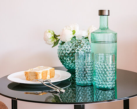 : Glastisch mit weißem Teller, auf dem ein Dessert und zwei Gabeln liegen, daneben zwei türkise Wassergläser und eine gefüllte Wasserflasche in derselben Farbe, sowie eine Vase mit weißen Blumen im Hintergrund.