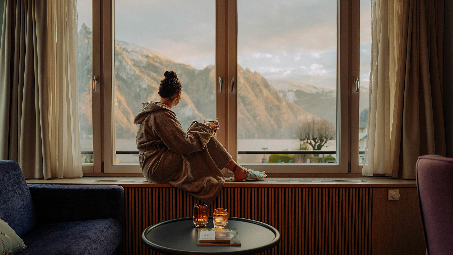  Frau im Bademantel sitzt auf der Fensterbank mit einem warmen Getränk und schaut auf den See und die schneebedeckten Berge.