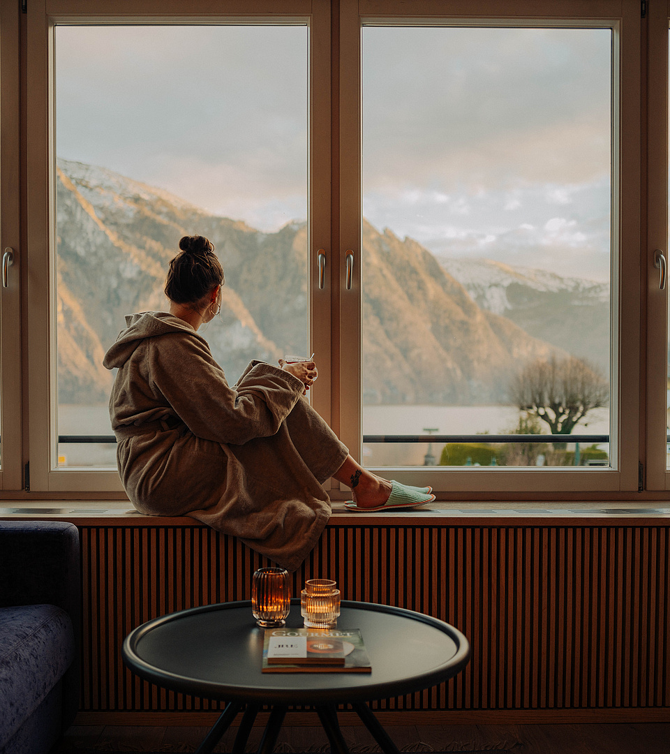  Frau im Bademantel sitzt auf der Fensterbank mit einem warmen Getränk und schaut auf den See und die schneebedeckten Berge.