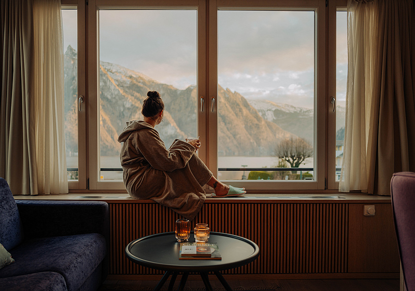  Frau im Bademantel sitzt auf der Fensterbank mit einem warmen Getränk und schaut auf den See und die schneebedeckten Berge.