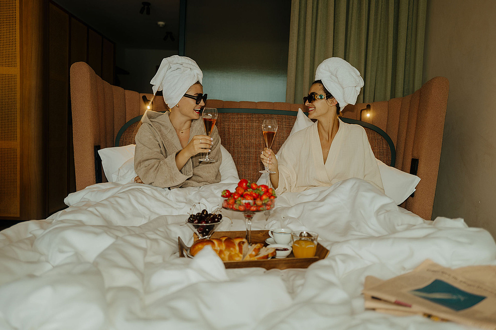 Zwei Frauen im Bademantel genießen ein Glas Sekt im Bett, mit einem Tablett voller Erdbeeren, Kirschen und Kuchen davor.