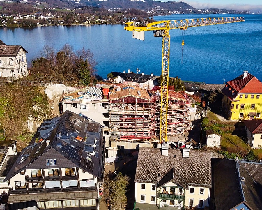 Drohnenaufnahme von Traunkirchen im Winter, mit der Baustelle des neuen Hotelzubaus, einem großen gelben Baukran, den Dächern der Häuser, dem See und den Bergen im Hintergrund.
