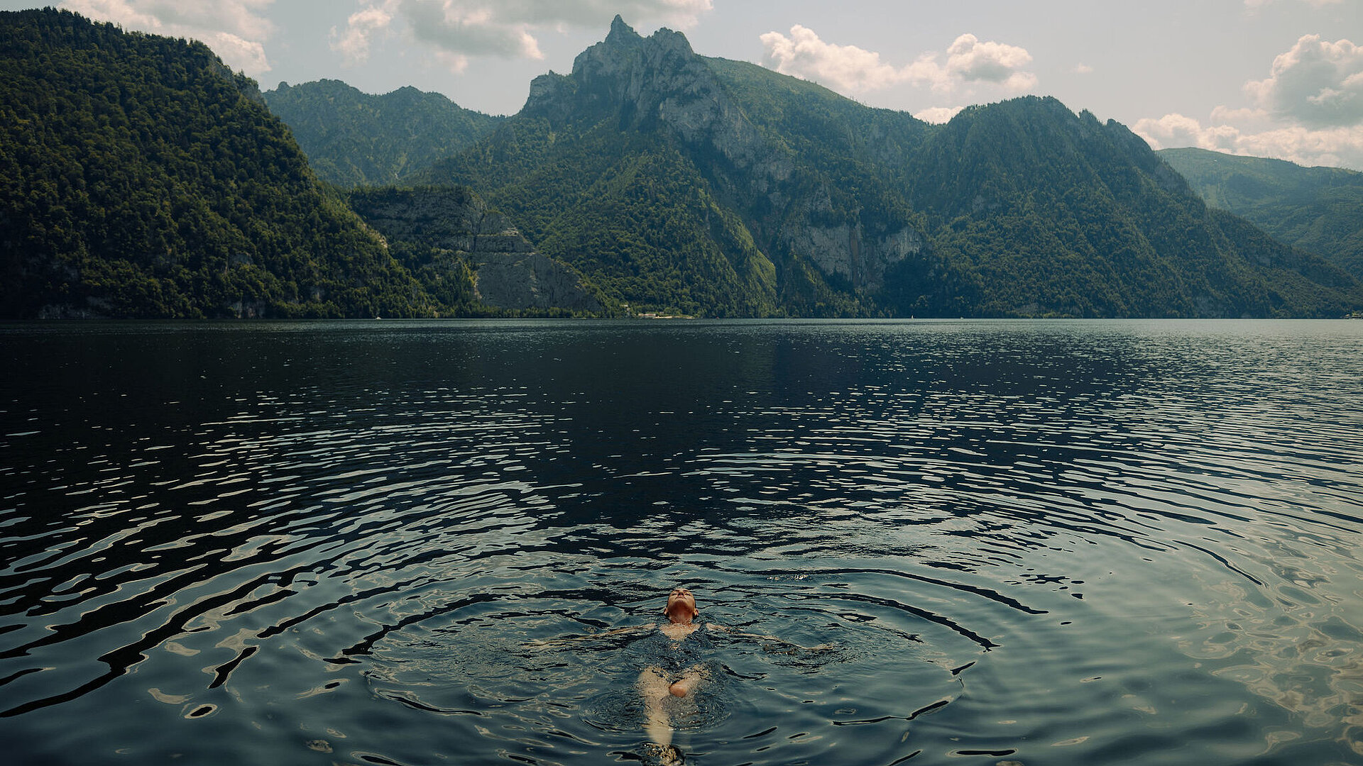 Frau schwimmt entspannt auf dem Rücken im See, grüne Berge im Hintergrund, Sommertag.