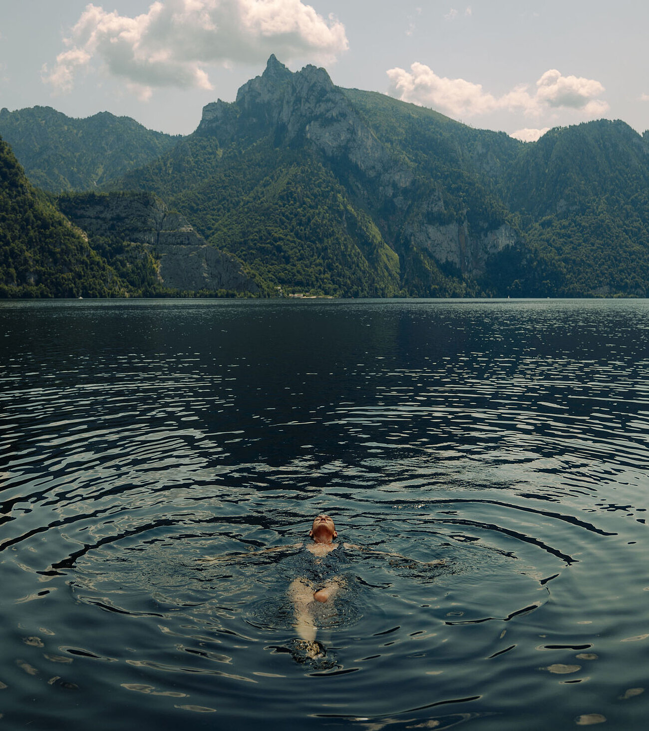 Frau schwimmt entspannt auf dem Rücken im See, grüne Berge im Hintergrund, Sommertag.