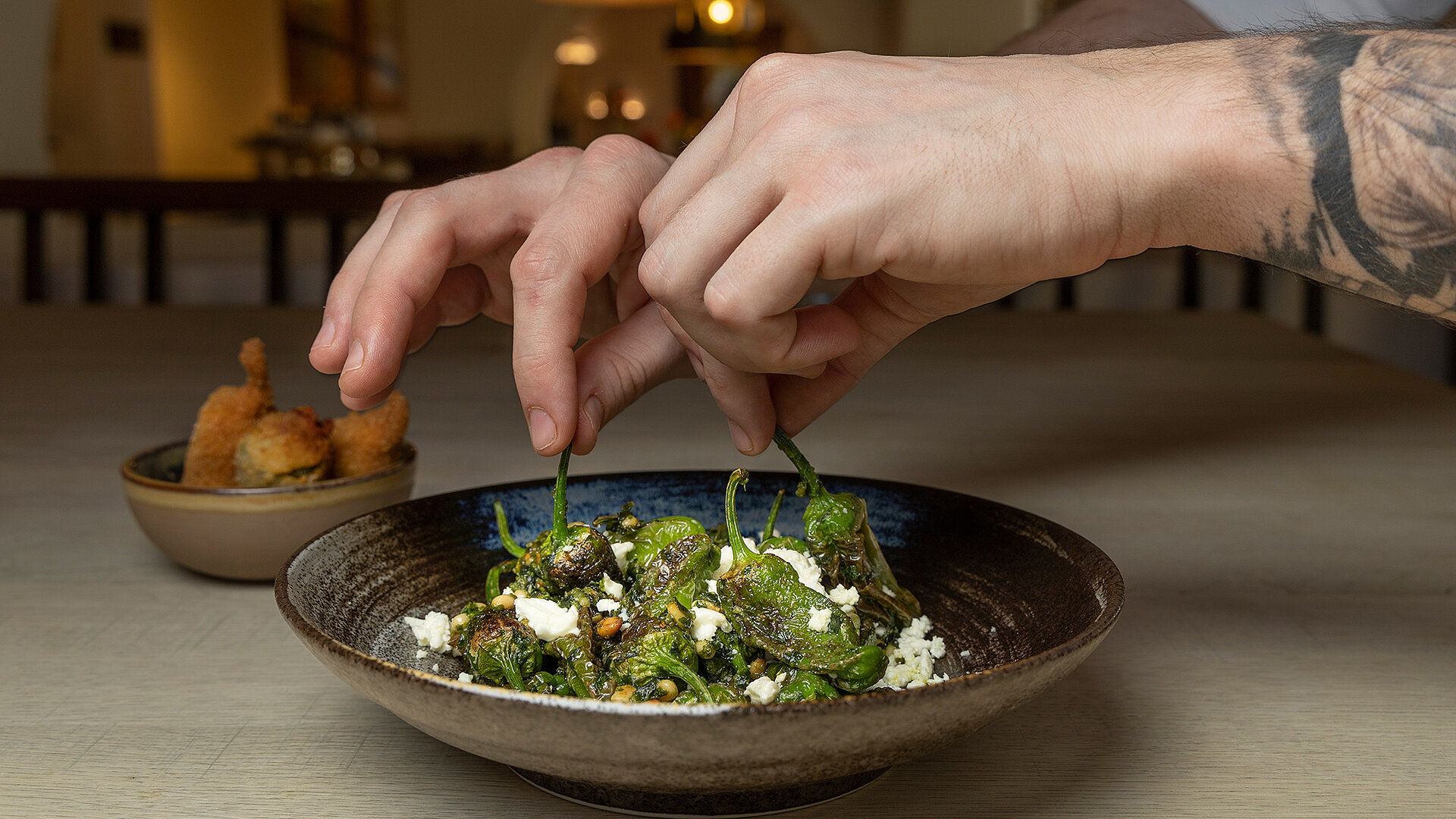 Hände beim Anrichten von Pimientos de Padrón im Restaurant Poststube, mit dem Restaurantinterieur im Hintergrund.