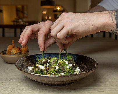 Hände beim Anrichten von Pimientos de Padrón im Restaurant Poststube, mit dem Restaurantinterieur im Hintergrund.
