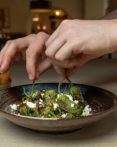 Hände beim Anrichten von Pimientos de Padrón im Restaurant Poststube, mit dem Restaurantinterieur im Hintergrund.