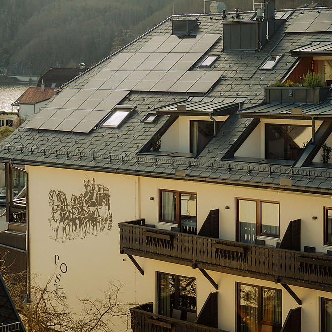 Seitenansicht des Hotels mit Balkonen und Logo an der Hauswand, im Hintergrund Berge und der Traunsee in der Ferne