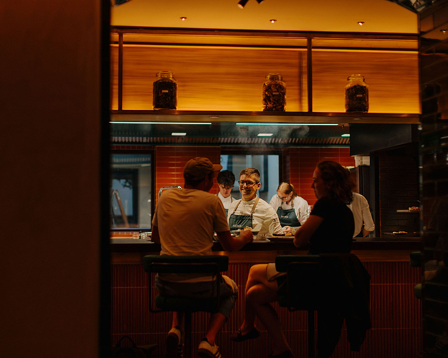 Offene Küche im Restaurant Beletage mit Köchen bei der Zubereitung. Gäste sitzen am Counter und beobachten das Geschehen