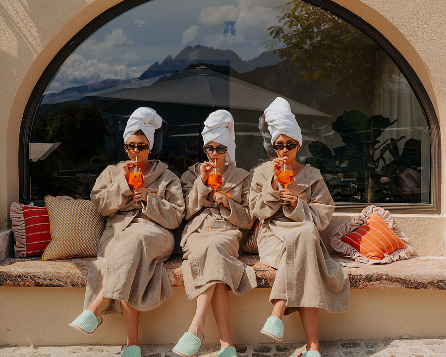 Drei Frauen im Bademantel und mit Handtüchern um den Kopf sitzen auf einer Steinbank vor dem Hotel und trinken orange Cocktails.
