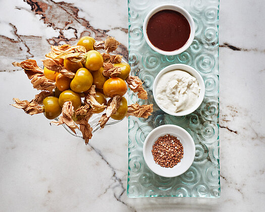 Drei kleine weiße Schälchen mit verschiedenen Saucen auf einem gläsernen Teller, daneben ein Glas mit Physalis, präsentiert auf einem weißen Marmortisch