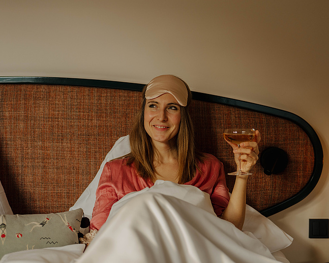 Frau mit Glas Sekt im Bett, lächelnd, in pinkem Morgenmantel, an gepolsterter Wand gelehnt, buntes Dekopolster sichtbar.