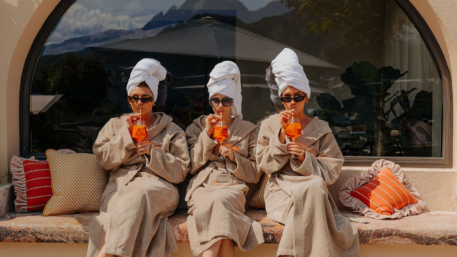 Drei Frauen im Bademantel und mit Handtüchern um den Kopf sitzen auf einer Steinbank vor dem Hotel und trinken orange Cocktails.