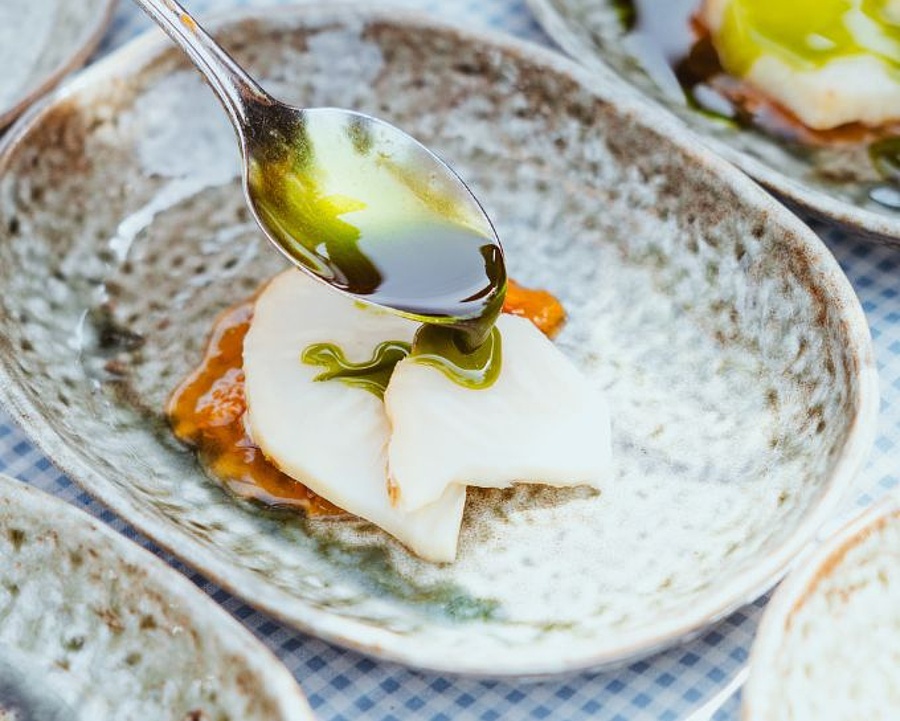 Weiß-blau karierte Tischdecke mit kleinen Schälchen voller Speisen. Im Vordergrund ein Fischgericht mit grünem Pesto und Löffel