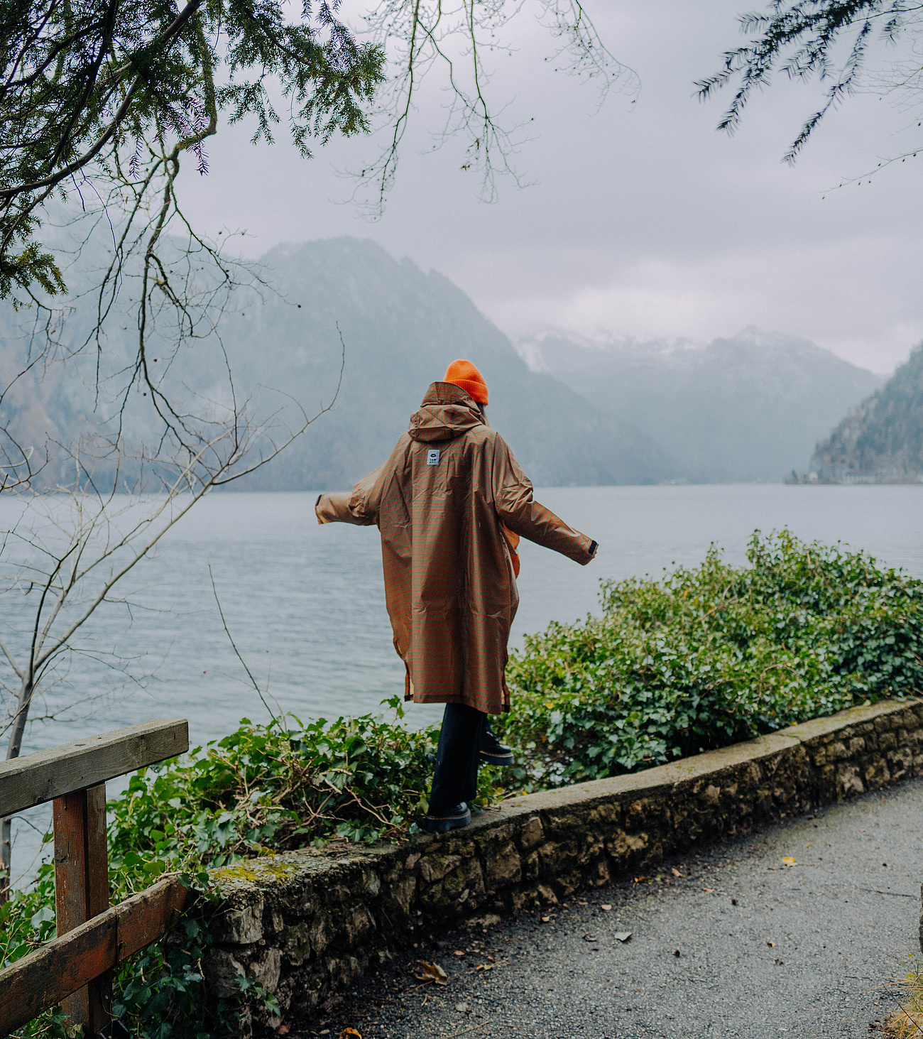 Person im braunen Regencape balanciert auf einer Steinmauer im Regen, im Hintergrund die Berge und der Traunsee