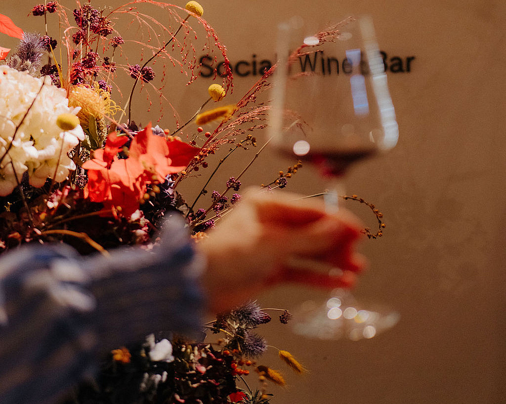 Hand mit Weinglas im Vordergrund, dahinter ein buntes Blumenarrangement und der Schriftzug „Social Wine Bar“ an der Wand