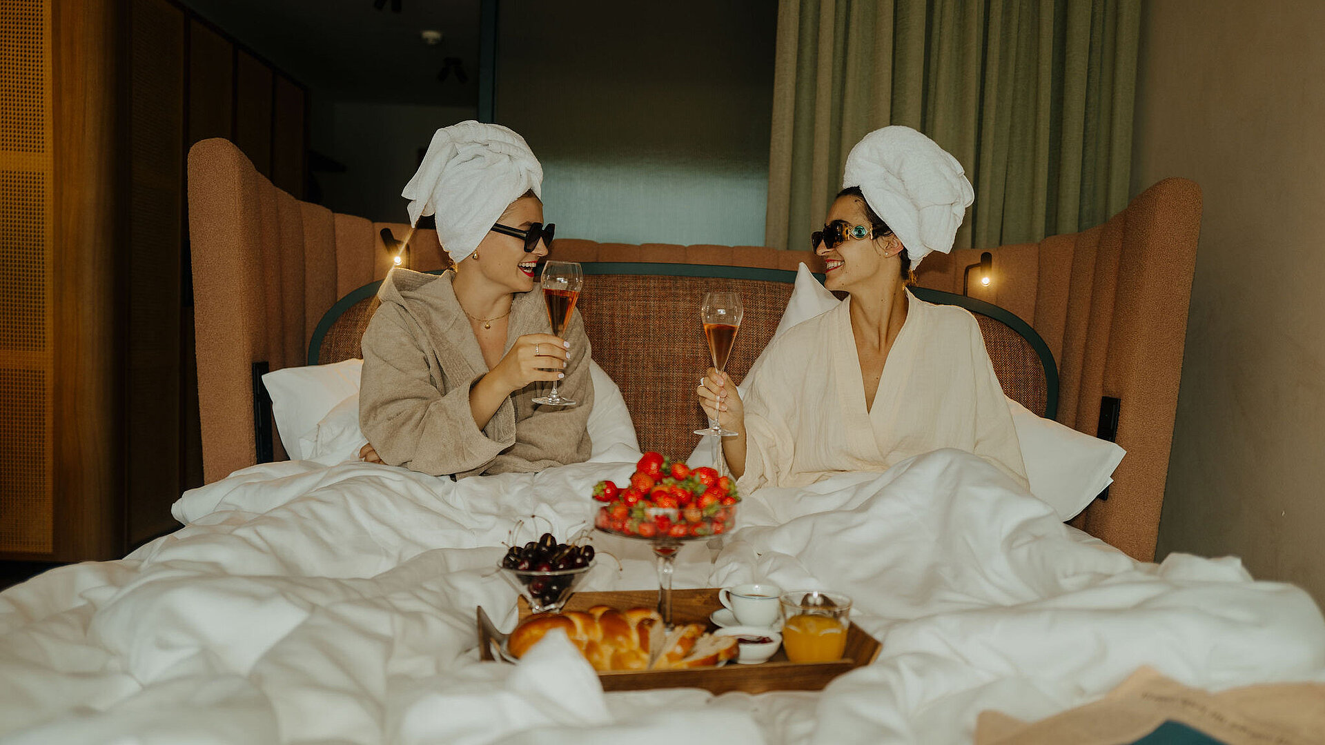Zwei Frauen im Bademantel genießen ein Glas Sekt im Bett, mit einem Tablett voller Erdbeeren, Kirschen und Kuchen davor.